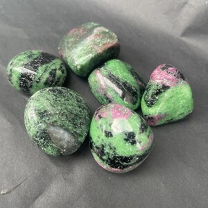 Könnte beinhalten: Eine Ansammlung von sechs polierten Trommelsteinen, die eine Mischung aus leuchtend grünem Zoisit, tiefschwarzer Hornblende und Flecken aus magentafarbenem Rubin aufweisen. Jeder Stein hat eine glatte, glänzende Oberfläche und unregelmäßige Formen.