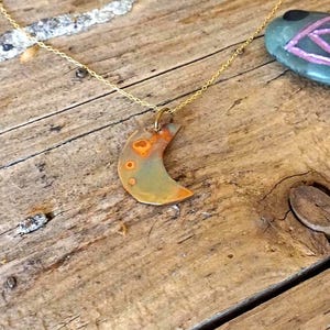 Może przedstawiać: Naszyjnik z wisiorkiem w kształcie półksiężyca z pomarańczowymi i zielonymi wzorami, na złotym łańcuszku. W tle widoczny jest kamień z różowym wzorem oka. Biżuteria leży na drewnianej powierzchni.