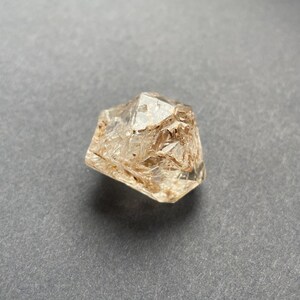 Könnte beinhalten: Ein klarer, facettierter Kristall mit einem hellbraunen Farbton. Der Kristall hat eine raue, strukturierte Oberfläche und ist vor einem grauen Hintergrund platziert. Der Edelstein scheint ein rohes, ungeschliffenes Mineral zu sein.