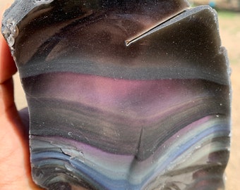 May include: A large, iridescent obsidian rock with a dark gray, purple, and blue color pattern. The rock has a smooth, polished surface and is shaped like a teardrop.