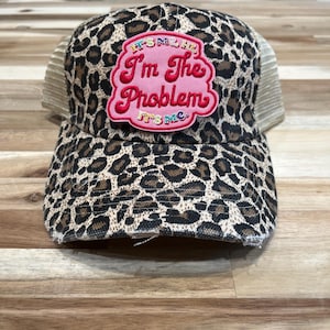May include: A trucker hat with a leopard print pattern in brown and black. The hat features a pink patch with the text "I'm The Problem" in red and white. The mesh back is tan.