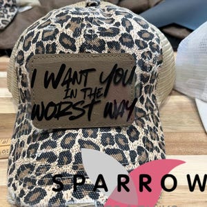 May include: A trucker hat with a leopard print pattern and a brown leather patch. The patch features the phrase "I WANT YOU IN THE WORST WAY" in black lettering. The hat has a mesh back and a distressed brim.