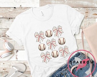 May include: White t-shirt with a baseball and bow design. The shirt features multiple baseballs with red stitching and bows in red and white.