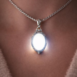 May include: A silver-toned oval pendant necklace with a glowing white center. The pendant has an ornate, filigree-style design around the edges. The chain is a delicate, linked style. The words "Glows Softly" are at the top.
