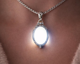 May include: An oval silver locket pendant with intricate filigree detailing hanging from a silver chain. The center of the locket emits a bright white light, and the text "Glows Softly" is printed in white above the pendant.