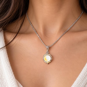 May include: A silver-toned box chain necklace featuring a circular pendant with an iridescent white and yellow center stone. The pendant is framed by intricate silver metalwork and small, sparkling yellow accent stones at the top and bottom.
