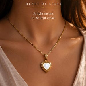 May include: A gold-colored heart-shaped pendant necklace with a white center stone. The necklace is worn on a person's neck. The image includes the text "COR LUMINIS HEART OF LIGHT" and "A light meant to be kept close."