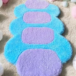Könnte beinhalten: Ein plüschiger, pfotenförmiger Teppich in Blau- und Lilatönen. Der Teppich hat einen hellblauen Außenrand und vier lila, rechteckige Akzente. Der Teppich besteht aus einem weichen, strukturierten Material.