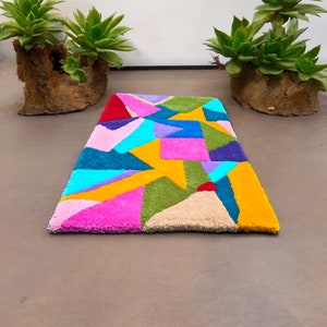 Könnte beinhalten: Ein bunter geometrischer Teppich in den Farben Rosa, Gelb, Grün, Blau und Rot. Der Teppich ist aus weichem, plüschigem Material gefertigt und hat ein modernes, abstraktes Design.