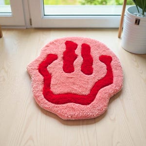 Könnte beinhalten: Ein rosa Teppich mit einem roten Smiley-Gesicht-Design. Der Teppich ist rund und hat ein weiches, strukturiertes Aussehen. Die roten Details des Gesichts, einschließlich der Augen und des Mundes, sind vor dem rosa Hintergrund deutlich definiert. Dieser dekorative Teppich eignet sich für ein Schlaf- oder Wohnzimmer.
