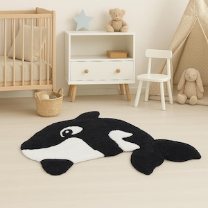 Puede incluir: Una alfombra con forma de orca en blanco y negro. La alfombra está hecha de un material suave y texturizado y presenta un diseño detallado del cuerpo de la orca, incluyendo sus distintivas marcas y aletas. La alfombra está colocada sobre un suelo de madera clara.