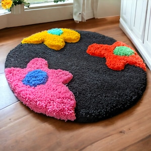 Könnte beinhalten: Ein runder schwarzer Teppich mit drei bunten Blumendesigns. Die Blumen sind rosa mit blauen Zentren, gelb mit grünen Zentren und orange mit grünen Zentren.