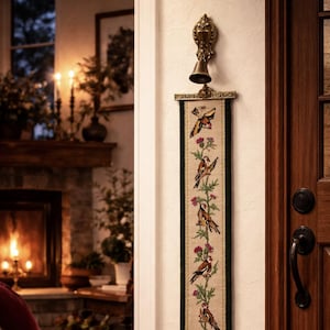 Puede incluir: Una campana de puerta decorativa con acabado de latón, montada sobre un tapiz largo y estrecho. El tapiz presenta pájaros bordados, flores y mariposas en tonos marrones, rosas y verdes. La campana cuelga cerca de una puerta de madera.