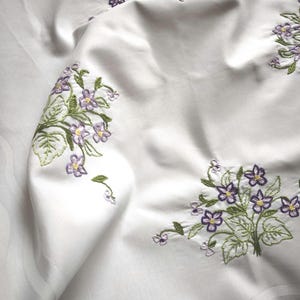 May include: A white fabric tablecloth featuring embroidered clusters of purple flowers with yellow centres and green leaves. The textile has a subtle, tone-on-tone woven striped pattern visible in the fabric weave throughout the surface.