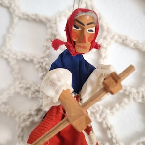 Può includere: Un ornamento a forma di bambola sospesa con volto in legno dipinto, fazzoletto rosso, top blu e gonna con motivo rosso, bianco e verde. La bambola ha braccia in legno e tiene in mano un piccolo attrezzo in legno a forma di croce, appesa a un filo.