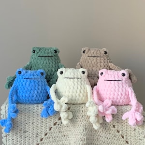 May include: Five crocheted frog plushies in different colors: green, blue, white, brown, and pink. The frogs are sitting on a crocheted blanket.