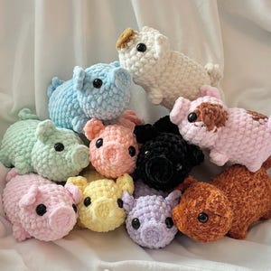May include: A collection of handmade crochet pig stuffed animals in various colors, including pink, blue, green, yellow, black, brown, and purple. Each pig has small black eyes and a curly tail. The pigs are arranged on a white, textured surface.