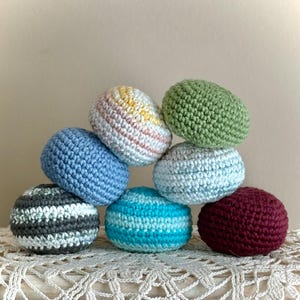 May include: A stack of eight crocheted spheres in various colors, including blue, green, white, and burgundy. Some spheres have striped patterns. The handmade items are arranged on a white lace surface.