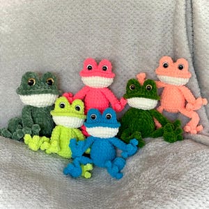 May include: A collection of six plush crochet frogs in various colors including dark green, lime green, bright pink, blue, forest green, and peach. Each frog features a white belly, large black plastic safety eyes, and articulated limbs.