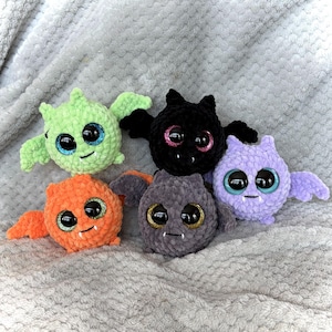 May include: A collection of six handmade crochet bat plushies in various colors. The bats have large, glittery eyes and small fangs. Colors include green, black, orange, gray, and purple. The plushies have small wings and smiling faces.