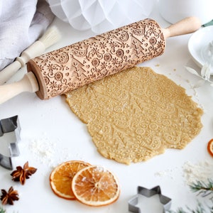 May include: A wooden rolling pin with a Christmas-themed design is used to roll out dough. The dough is imprinted with a pattern of snowflakes, trees, and birds.