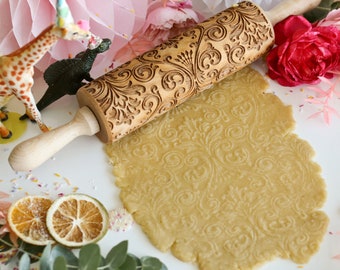 May include: A wooden rolling pin with a carved floral design is used to roll out dough, creating a decorative pattern on the surface. The dough is a light brown color and is surrounded by sprinkles and other decorative elements.