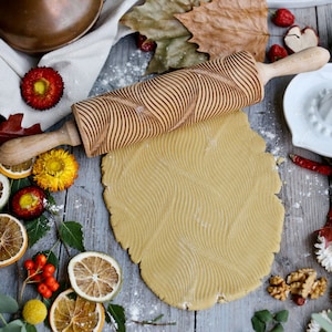 May include: A wooden rolling pin with a wavy pattern is used to roll out dough on a wooden surface. The dough has a wavy pattern imprinted on it.