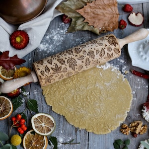 Pode incluir: Um rolo de massa de madeira com um padrão de folhas esculpidas é usado para abrir a massa. A massa é impressa com o padrão das folhas. O rolo está sobre uma superfície de madeira com decorações de outono.