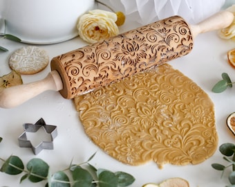 May include: A wooden rolling pin with an intricate floral design carved into the surface. The rolling pin is being used to roll out dough, which is imprinted with the design. The dough is a light brown color and is surrounded by dried apple slices and eucalyptus leaves.