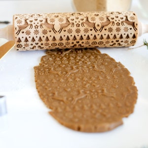 Puede incluir: Un rodillo de madera con un diseño navideño con renos, copos de nieve y árboles. El rodillo se utiliza para crear una impresión en una masa de galletas marrón.