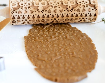 Pode incluir: Um rolo de massa de madeira com um design de Natal com renas, flocos de neve e árvores. O rolo é usado para criar uma impressão em uma massa de biscoito marrom.