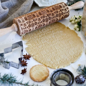 May include: A wooden rolling pin with a carved design of houses, trees, and stars. The rolling pin is used to create a patterned design in a sheet of dough. The dough is on a white surface with a cookie cutter and star anise.