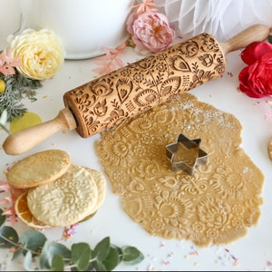 Op de afbeelding: Een houten deegroller met een bloemenpatroon, naast uitgerold deeg met hetzelfde ontwerp. Een stervormige koekjesvorm en reliëfkoekjes suggereren bakken. De deegroller is ongeveer 38 cm lang.