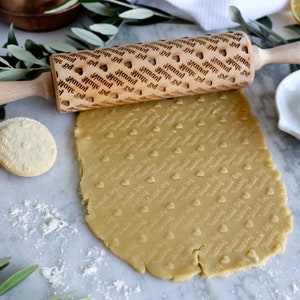 May include: A wooden rolling pin with a pattern of hearts and the text "Made by Nanny" repeated across the surface. The rolling pin is used to imprint the pattern onto a sheet of cookie dough.