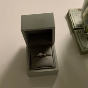 Könnte beinhalten: Eine graue Ringbox mit einem silbernen Ring im Inneren. Der Ring hat einen kleinen Diamanten in der Mitte. Die Box steht auf einer weißen Oberfläche. Rechts neben der Box liegen Stapel von US-Dollar-Scheinen.