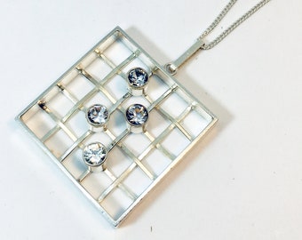 May include: Silver pendant necklace with a geometric, grid-like design. The square pendant has a lattice pattern and four clear, round gemstones. The necklace chain is silver and delicate. The pendant is approximately 3 cm wide.