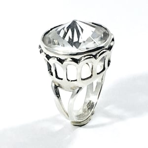 Peut inclure: Une bague en argent avec une grande pierre précieuse facetée transparente sertie dans une monture décorative ajourée.