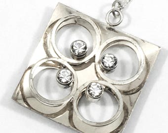 May include: A silver pendant necklace with a square-shaped design featuring four clear rhinestones set within concentric circles. The pendant is attached to a silver chain.