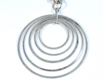 Pode incluir: Colar prateado com um pendente de seis círculos concêntricos graduados. Os círculos são ligados por um pequeno laço a uma corrente delicada. A joia está sobre um fundo branco.