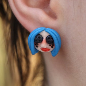 May include: A pair of blue and black polymer clay earrings featuring a stylized cartoon face with blue hair and black eyes.