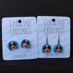 May include: Two pairs of Coraline earrings, one pair is stud earrings and the other pair is dangle earrings. Both pairs feature a blue-haired doll with button eyes. The cards say "Coraline Earrings" and "By Owlieart".