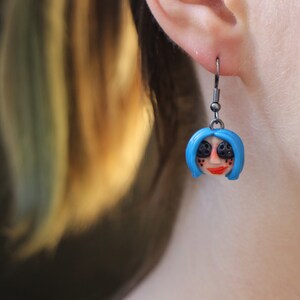 May include: A pair of earrings featuring a blue-haired doll face with black eyes and red lips. The earrings are made of polymer clay and have silver hooks.