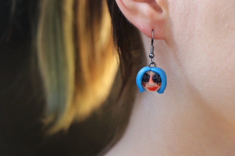 May include: A pair of earrings featuring a blue-haired doll face with black eyes and red lips. The earrings are made of polymer clay and have silver hooks.