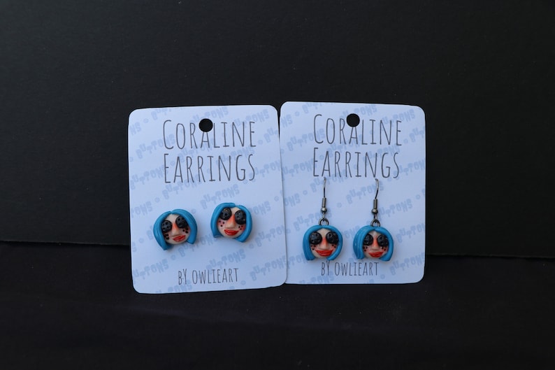 May include: Two pairs of Coraline earrings, one pair is stud earrings and the other pair is dangle earrings. Both pairs feature a blue-haired doll with button eyes. The cards say "Coraline Earrings" and "By Owlieart".