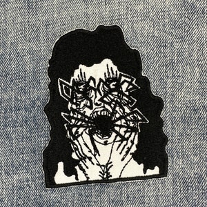 May include: Black and white embroidered patch of a screaming face with multiple eyes and hands around the face.