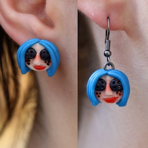 May include: A pair of earrings featuring a blue-haired doll head with black eyes and a red mouth. The earrings are made of polymer clay and have silver hooks.