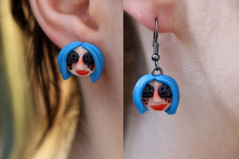 May include: A pair of earrings featuring a blue-haired doll head with black eyes and a red mouth. The earrings are made of polymer clay and have silver hooks.