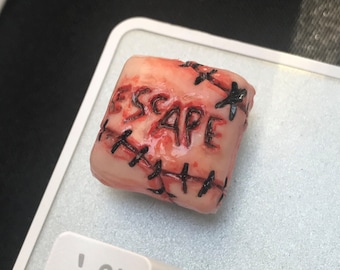May include: A flesh-colored keycap with the word "ESCAPE" written in black lettering. The keycap is decorated with black stitching, resembling stitches on a wound.