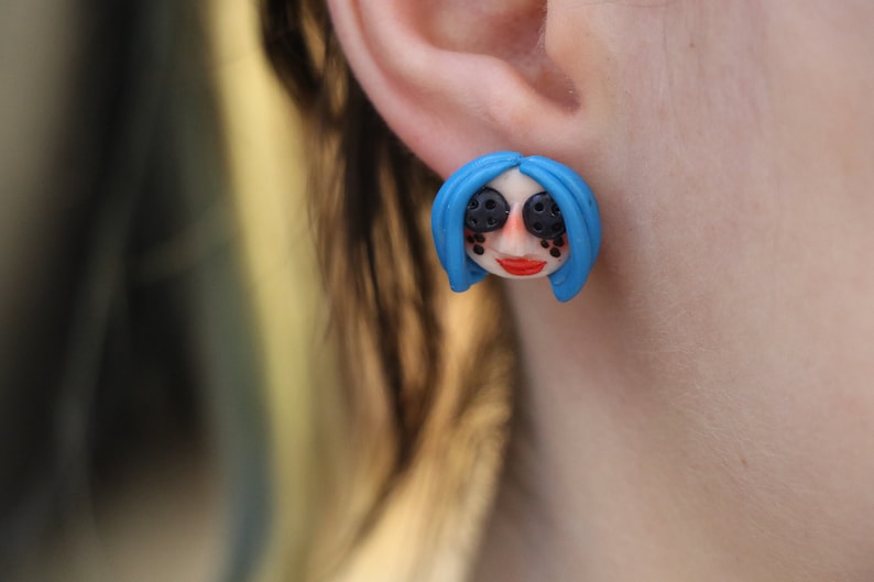 May include: A pair of blue and black polymer clay earrings featuring a stylized cartoon face with blue hair and black eyes.