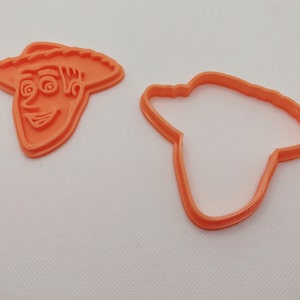 Può includere: Due formine per biscotti in plastica arancione. Una formina ha la forma del volto di un personaggio dei cartoni animati, con un cappello e un'espressione sorridente. L'altra formina è il contorno del cappello. Le formine sono su una superficie bianca.